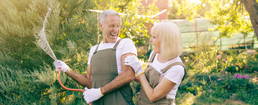 A cheerful couple shares a light moment while watering plants in their garden. Bright sunlight filters through green leaves, highlighting their playful interaction and love for gardening. - Powered by Adobe