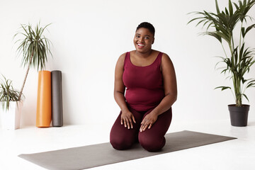 A cheerful overweight black lady yoga instructor greets her students while sitting on a fitness mat in a bright yoga studio. Her warm smile creates an inviting atmosphere for the morning workout.