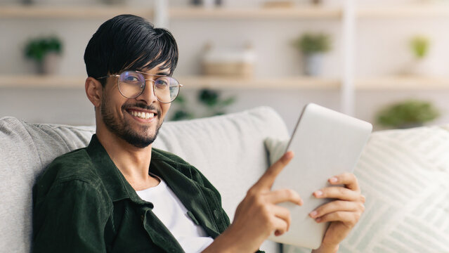 A young man with glasses sits on a cozy couch, smiling as he uses a tablet. The bright and welcoming living room features plants on shelves in the background.