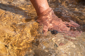 Close-up of a woman's hand touching a warm, smooth beach stone washed by clear, flowing seawater in summer