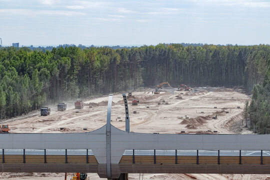 Large-scale highway construction site clearing a forest area, viewed over noise barrier. Heavy machinery performs earthworks and infrastructure development, highlighting human impact and urbanization.