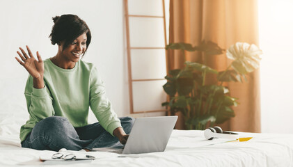 A woman sits on a bed in a bright room, smiling and waving at her laptop. She enjoys her time while wearing a green sweater, with plants and sunlight creating a welcoming atmosphere.
