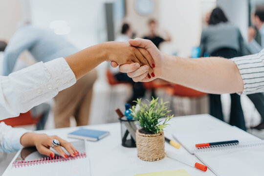 Two coworkers from diverse backgrounds shake hands over a shared desk in a modern office, symbolizing collaboration, teamwork, and partnership.