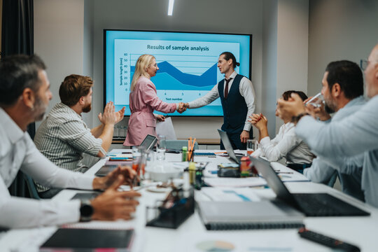 A diverse group of professionals in a modern conference room celebrate a successful handshake following a data analysis presentation.