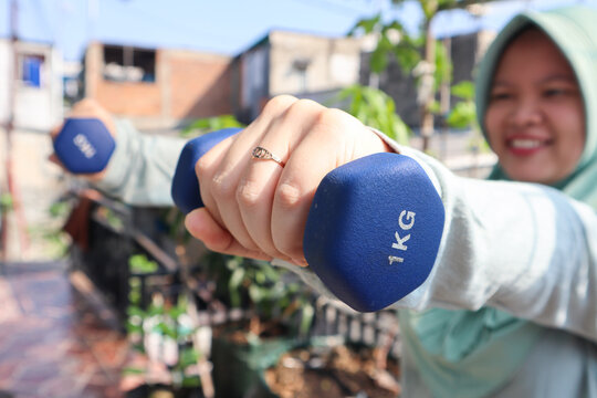 Young asian muslim woman, weightlifting and exercise with dumbbells for training, workout or muscle gain on outdoor balcony. Young female person with small barbells for sport, fitness or health. 