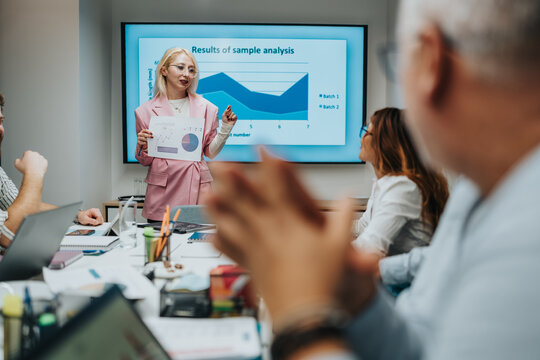 A presenter in a pink suit leads a business meeting, displaying charts on a screen while colleagues work on laptops. The focused team discusses data, insights, and results in a modern office. - Powered by Adobe