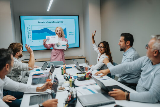 A diverse team gathers around a long table for a business meeting, listening to a presenter showing charts on a screen. Laptops, notes, and collaborative energy reflect a data-driven workplace.