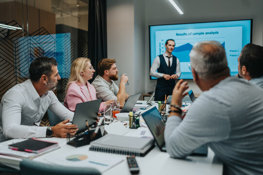 A group of professionals gathers around a conference table as a presenter explains a data chart shown on a large screen. Laptops, notebooks, and glasses indicate a focused business discussion.