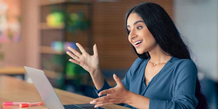 A woman is animatedly participating in a virtual meeting, using gestures and expressions to convey her enthusiasm. She sits at a desk, working on a laptop in a vibrant office environment.