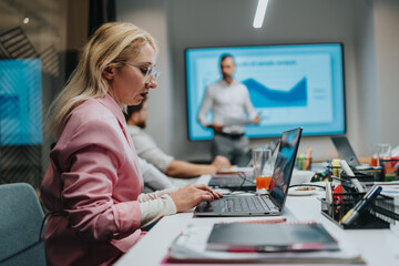 A focused professional woman in a pink blazer types on a laptop while colleagues discuss a charted presentation on a large screen in a modern office setting.