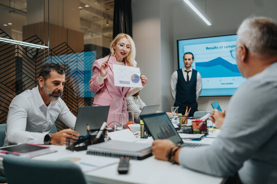 A diverse team gathers for a business meeting as a confident presenter shares charts and data on a large screen. Laptops, notebooks, and coffee cups fill a collaborative workspace.