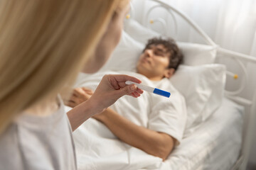 women holding pregnancy test while looking at sleeping boyfriend in bed. Concept of unexpected news, pregnancy result, family planning, relationship decision, surprise, emotional moment.