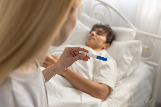 women holding pregnancy test while looking at sleeping boyfriend in bed. Concept of unexpected news, pregnancy result, family planning, relationship decision, surprise, emotional moment.