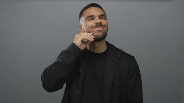Young hispanic man with hand on chin and head tilted upward in grey studio showing pensive pose; idea reflection.