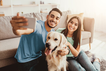 A young couple happily poses for a selfie on their couch with a golden retriever. Sunlight brightens the cozy living room, creating a joyful atmosphere for the moment.
