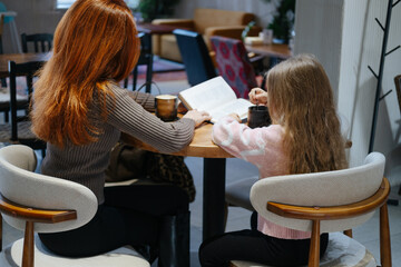 Pretty caucasian mother and her daughter sitting in the cafe and drinking cacao or coffee. Reading and looking at the book. Slow motion of cute young woman with her kid