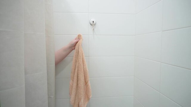 The hand takes a terry towel from the hook into the shower. White tiles and shower curtain, bright bathroom, close-up.
