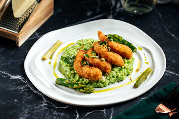 Creamy green risotto with golden fried shrimp and asparagus served on white plate over dark table