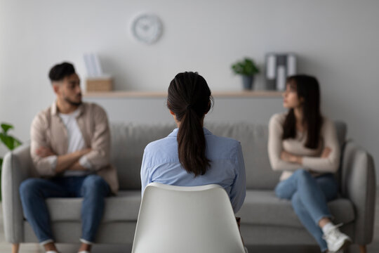 In a therapy session, a female psychologist is assisting a quarreling middle eastern couple. The scene captures the back view of the psychologist as she guides the couple toward resolution.