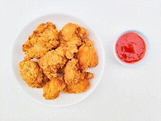 Fried chicken nuggets in a white plate and sauce in a small white bowl on a white background.