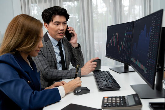 Smiling Asian businessman calling to broker dynamic stock exchange database with woman business partner pointing to screens comparing profit value online application program at home office. Infobahn.