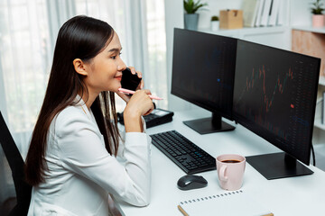 Smiling young beautiful Asian businesswoman calling to broker on smartphone to analyze exchange stock market graph on pc screen at modern office. Concept of investing high profit in wealth. Stratagem.