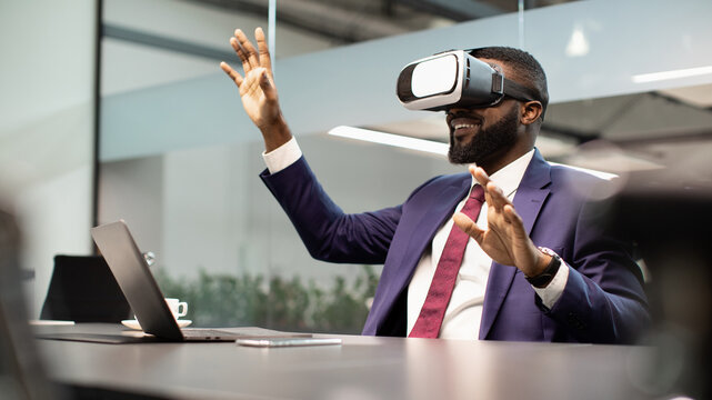 Modern technologies in business, virtual reality for enterprise concept. Excited black businessman sitting at workdesk in front of laptop, using VR glasses, touching copy space, modern office interior
