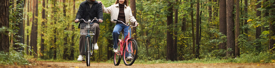 Obraz premium Header of Young adult Caucasian man and young adult Caucasian woman riding bicycles together on forest path, smiling and enjoying outdoor activity, surrounded by tall trees and greenery