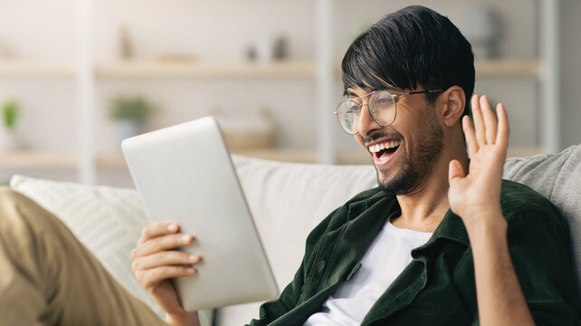 A man with glasses is smiling and waving while having a video call. He sits comfortably on a couch in a modern living room, using a tablet to connect with someone remotely.