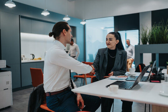Two professionals greet with a handshake in a bright office setting, surrounded by laptops, coffee, and teamwork.