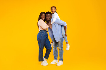 A joyful African American girl is having fun on her father's back while her mother smiles beside them. The family is posing together in a cheerful atmosphere with an orange background.