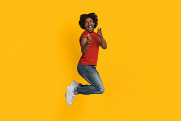 A young man in a red shirt and blue jeans jumps in excitement, smiling widely. He poses playfully with finger guns against a lively yellow background, showcasing pure joy and energy.