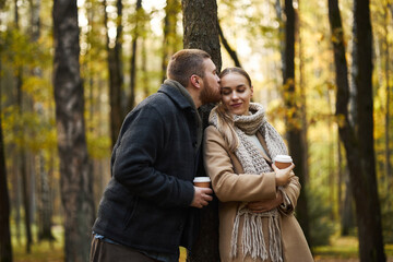 Caucasian young adult man kissing forehead of girlfriend holding coffee cup, standing together in autumn forest, both wearing warm clothing and appearing affectionate