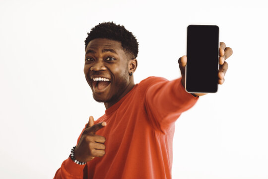 A cheerful young man wearing a red sweater stands indoors, pointing and smiling while holding a smartphone. His joyful expression conveys excitement and enthusiasm.