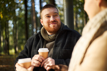 Caucasian young adult man smiling while holding two takeaway coffee cups, sitting outdoors in park with blurred in foreground, engaging in friendly conversation with girlfriend