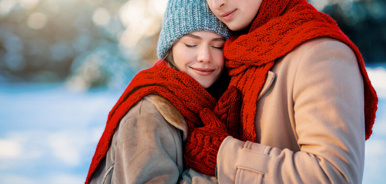 A young couple stands close together in a snowy landscape, sharing a warm embrace. They wear thick coats and bright red scarves, radiating warmth amidst the cold winter setting.
