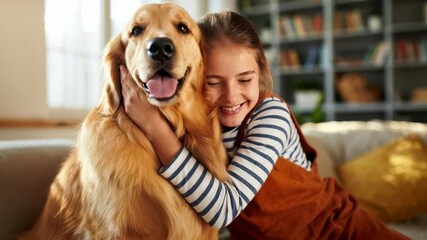 A happy little girl hugs a golden retriever dog on the sofa at home, demonstrating love and friendship for pets.