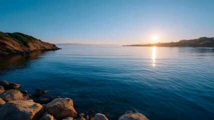 Calm ocean bay at sunrise with a rocky shore and distant coastline under a clear blue sky