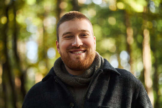 Portrait of young Caucasian man smiling outdoors with short hair and beard, looking directly at camera, standing in forest setting with blurred green background during nature walk