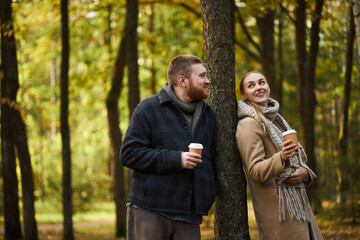 Caucasian young man and Caucasian young woman standing in autumn forest holding takeaway coffee...