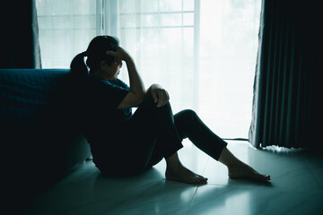 A lonely woman sits on the floor by window with hand on head in dark room. Concept of depression, sadness, anxiety, stress, mental health problem, loneliness and emotional struggle in life.