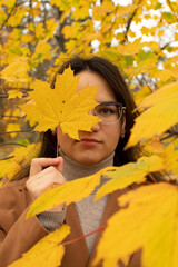 Portrait of a young attractive woman in eyeglasses covering her face with a yellow maple leaf	