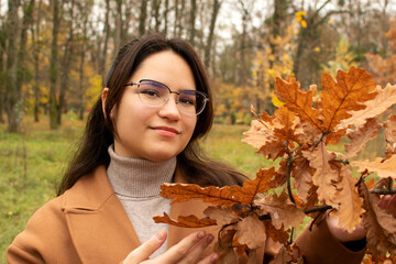 Portrait of a young attractive woman wearing a brown coat standing under an oak tree in autumn park	