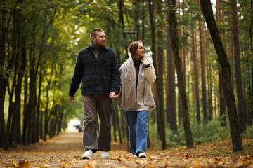 Fototapeta premium Caucasian young adult man and Caucasian young adult woman walking together holding hands in forest park, both looking at each other and smiling, autumn leaves covering ground