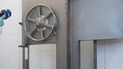 A person in a lab coat and gloves washes down an industrial meat cutting machine with a spray hose in what looks like a butcher shop or food processing plant during the day.