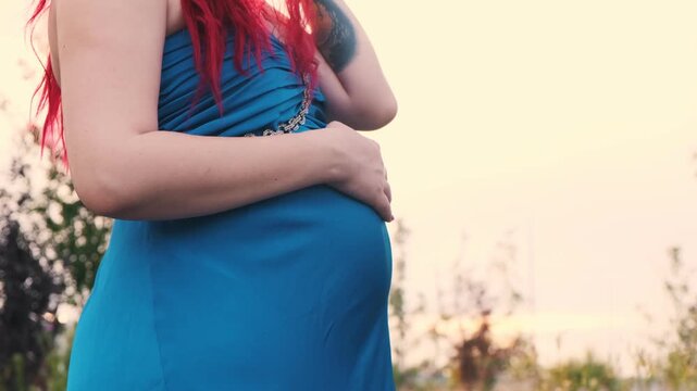 A pregnant woman in a blue dress gently strokes her round belly with her hand, embracing the moment of anticipation in a public urban park at sunset
