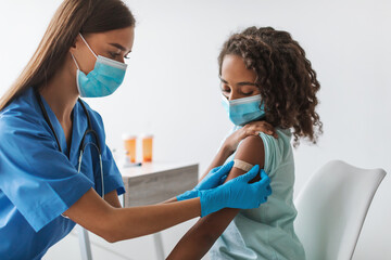 In a clinic, a medical worker is giving a vaccine to a Black pre-adolescent girl. The girl wears a face mask and receives an adhesive bandage on her arm after the shot.