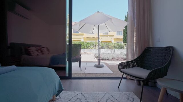 Smooth camera gliding through modern bedroom interior, gradually revealing sun drenched patio with relaxing outdoor seating arrangement and parasol, highlighting indoor outdoor living connection