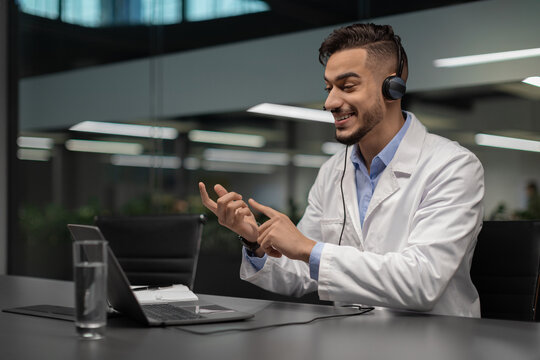 A handsome young middle eastern scientist is sitting in a modern laboratory. He is wearing workwear and using a laptop for a video chat with colleagues, smiling and gesturing enthusiastically. - Powered by Adobe