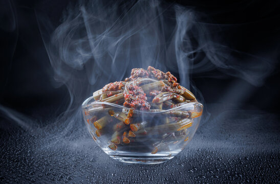 Smoke curls around a clear glass bowl of bracken fern salad with spices. Fern salad on a black background.